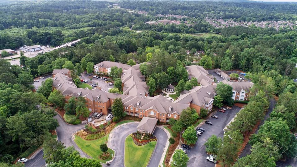 Aerial view of Galleria Woods senior living facility surrounded by dense green trees. The large, multi-wing building has a circular driveway at the entrance with landscaped greenery and parking areas around the building. The facility is nestled in a heavily wooded area with other buildings visible in the distance.