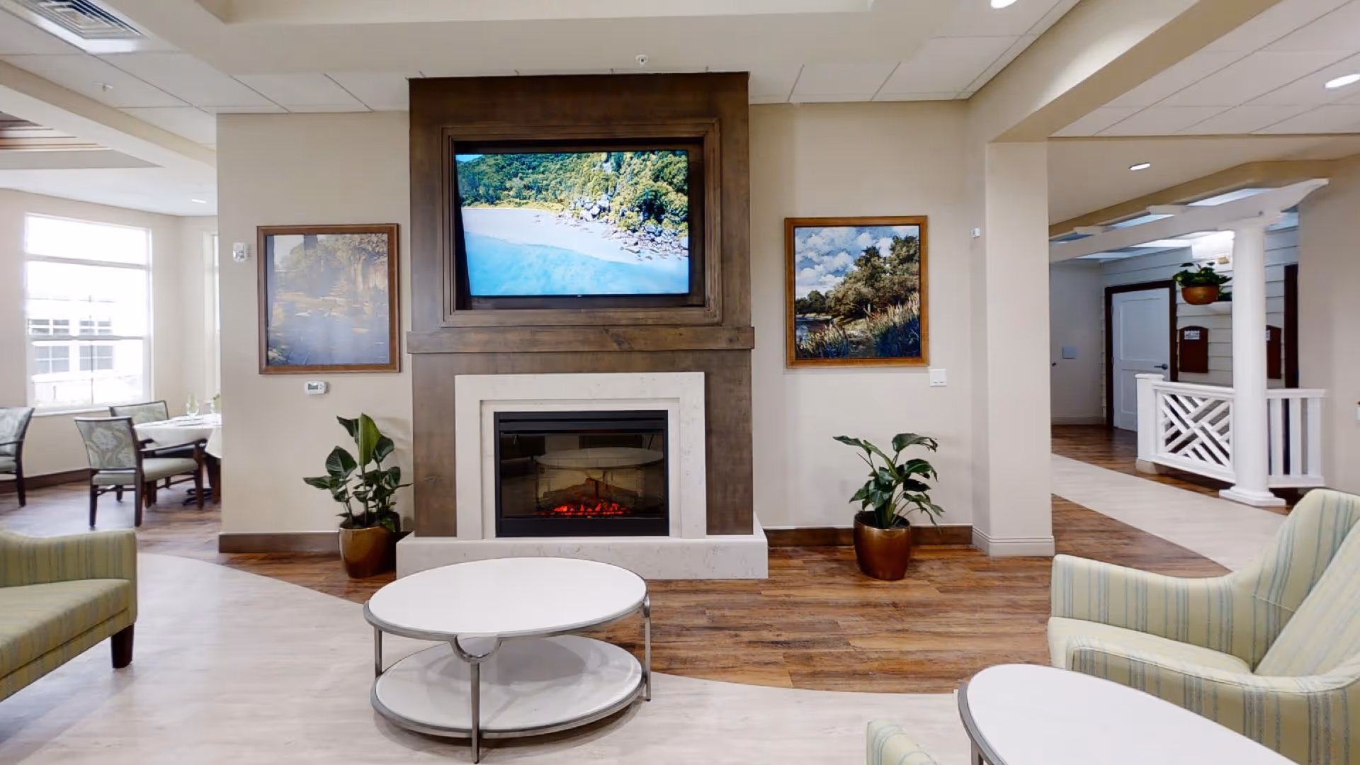 A cozy living room area with a modern electric fireplace set in a wooden frame. Above the fireplace is a large flat-screen TV displaying a beach scene. On either side of the fireplace are framed landscape paintings and potted plants. The room features wood flooring, light-colored walls, and comfortable striped armchairs. In the background, there is a dining area with tables and chairs near large windows.