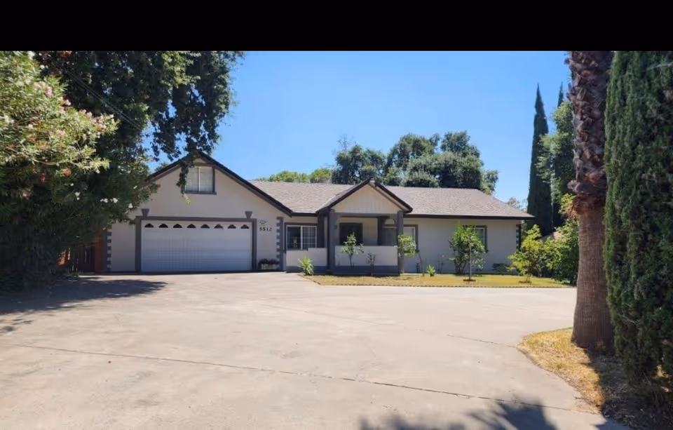 Single-story residential building with a two-car garage, driveway, and trees in the front yard.