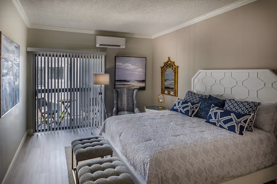 A well-decorated bedroom with a large white bed featuring blue and white starfish-themed pillows. There are two tufted benches at the foot of the bed. A patterned armchair and a floor lamp stand near a sliding glass door with vertical blinds leading to a small outdoor patio with chairs and a table. The walls are adorned with a large painting, a decorative mirror, and the room has light-colored flooring.