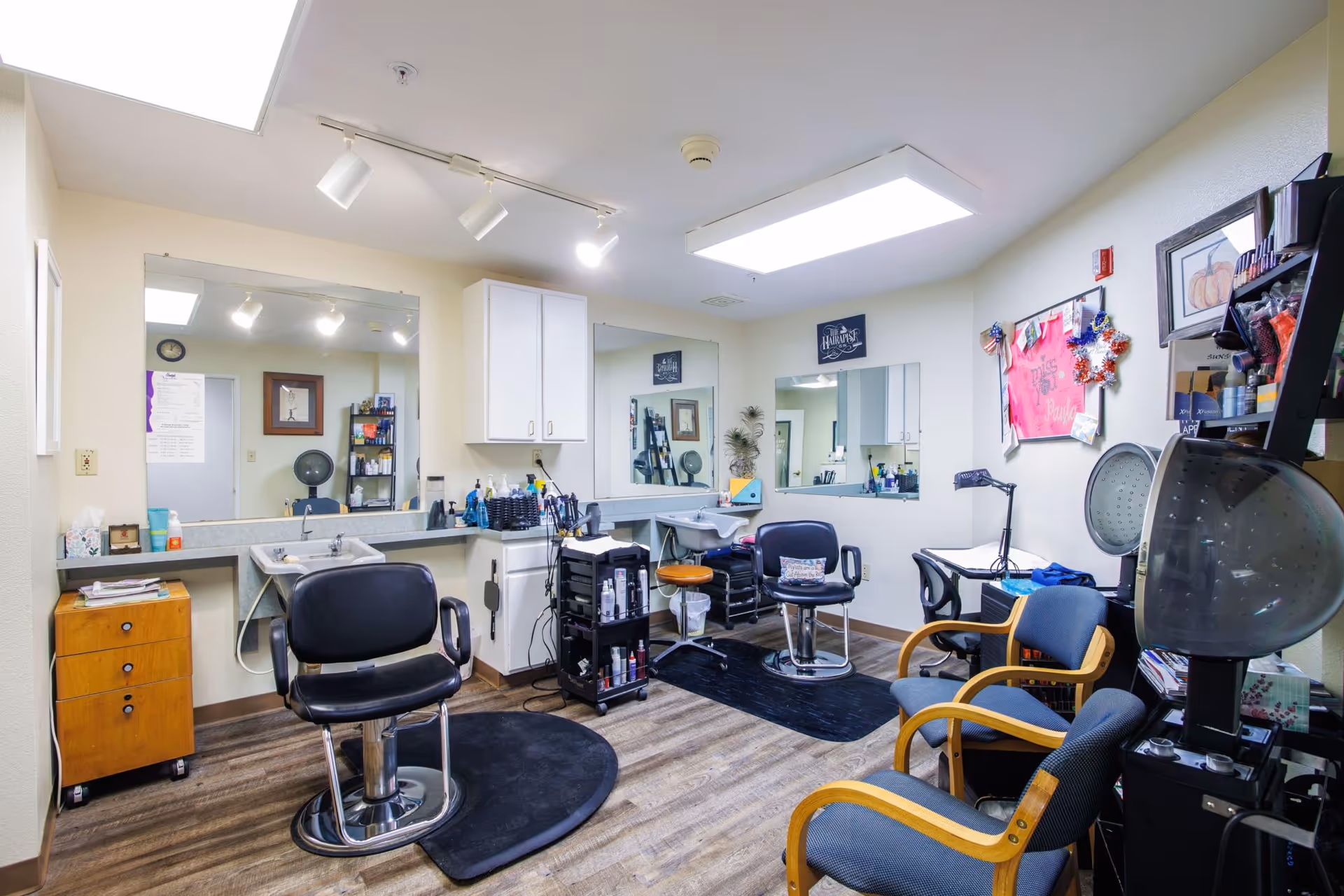 Hair salon room with styling chairs, sinks, mirrors, hairdryers, and shelves of hair products in an assisted living facility.