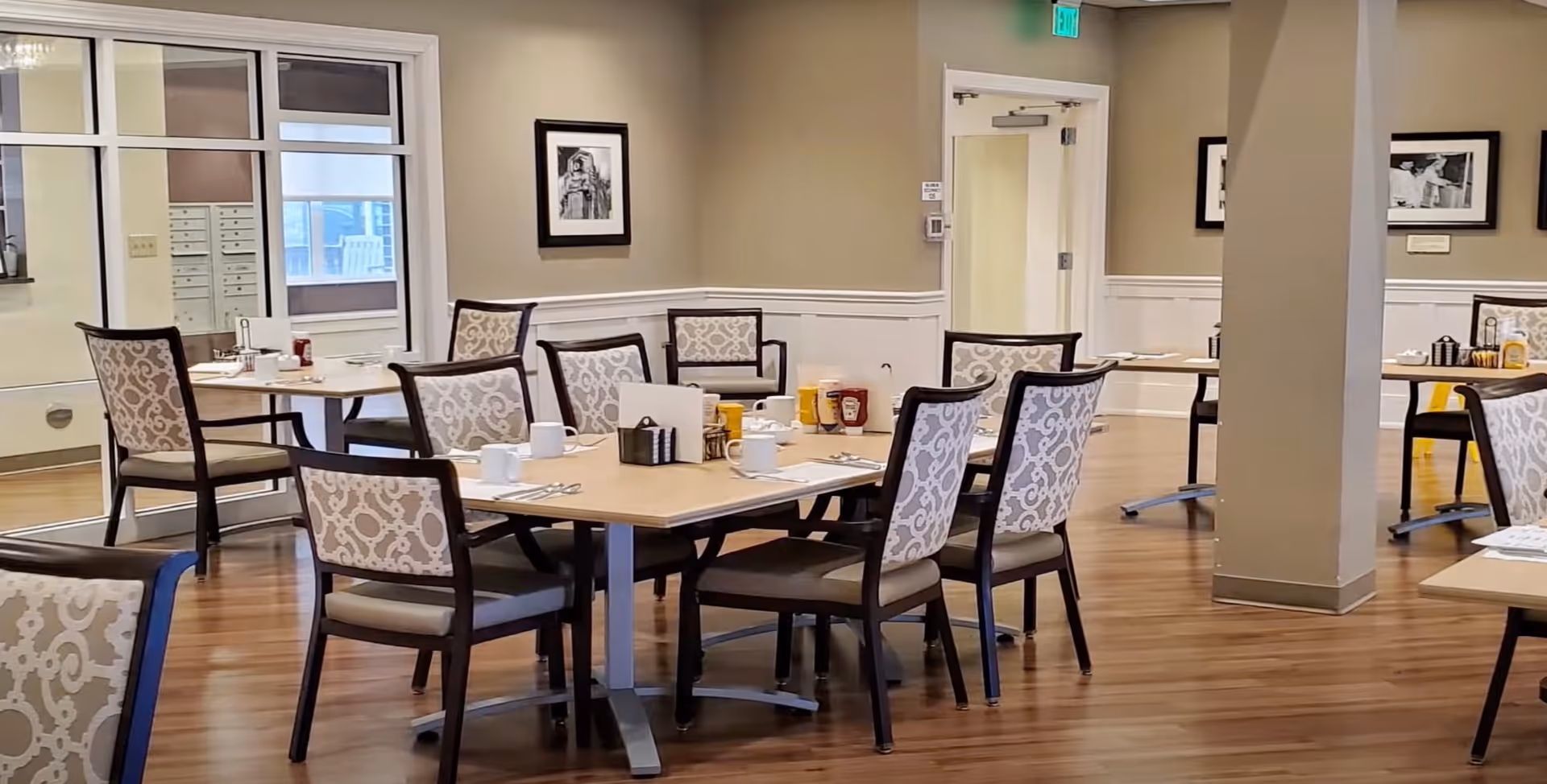 Dining room with multiple tables and patterned chairs set for meals in a senior living facility.