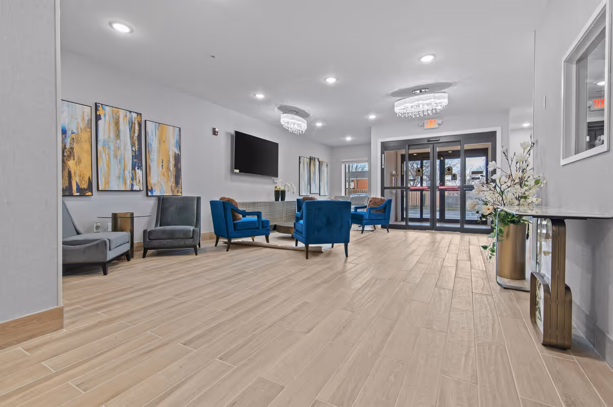 A modern and spacious lobby area with light wood flooring, blue and gray upholstered chairs arranged around a circular rug, abstract artwork on the walls, a flat-screen TV mounted on the wall, and glass double doors leading outside. There are decorative plants and a console table on the right side.