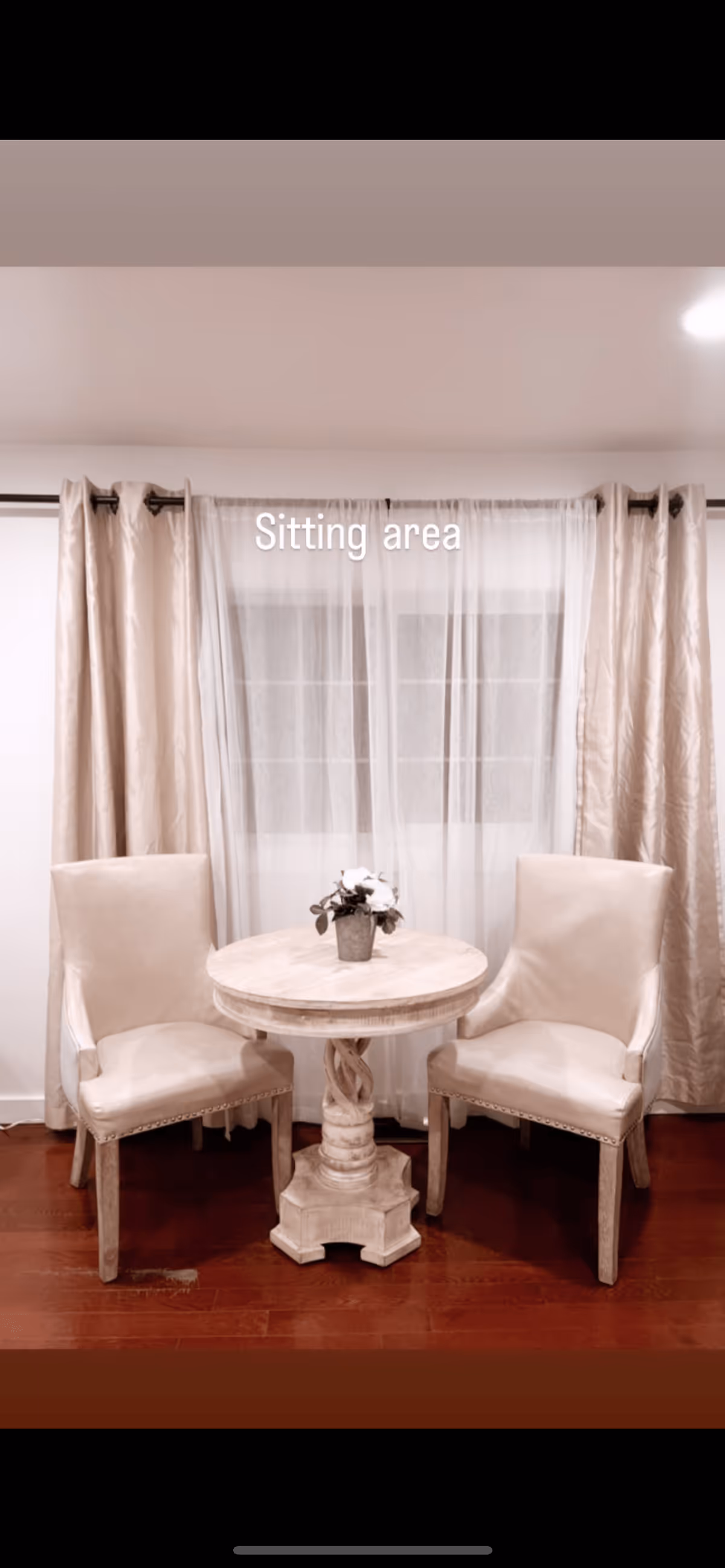 A small sitting area with two beige upholstered chairs and a round wooden table with a decorative base. A small potted plant is placed on the table. Behind the sitting area is a window with sheer white curtains and beige drapes on either side.