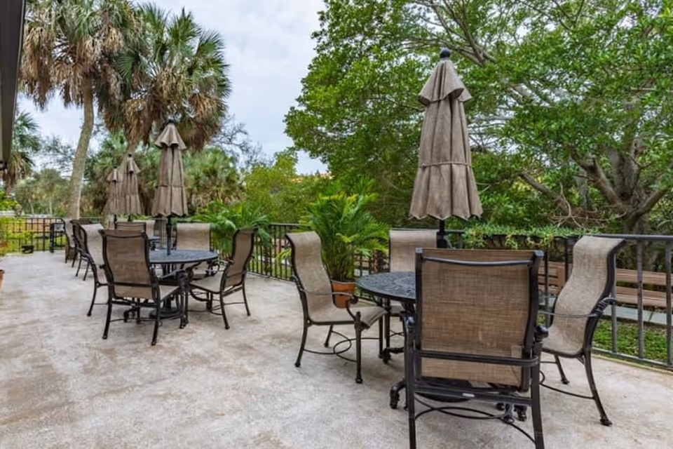 Outdoor patio area with multiple round metal tables and beige mesh chairs. Each table has a closed beige umbrella. The patio is surrounded by lush green trees and palm trees, creating a serene and shaded environment.