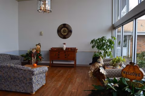 Bright lounge with patterned sofas, a wooden console table under a wall clock, potted plants, and floor-to-ceiling windows.