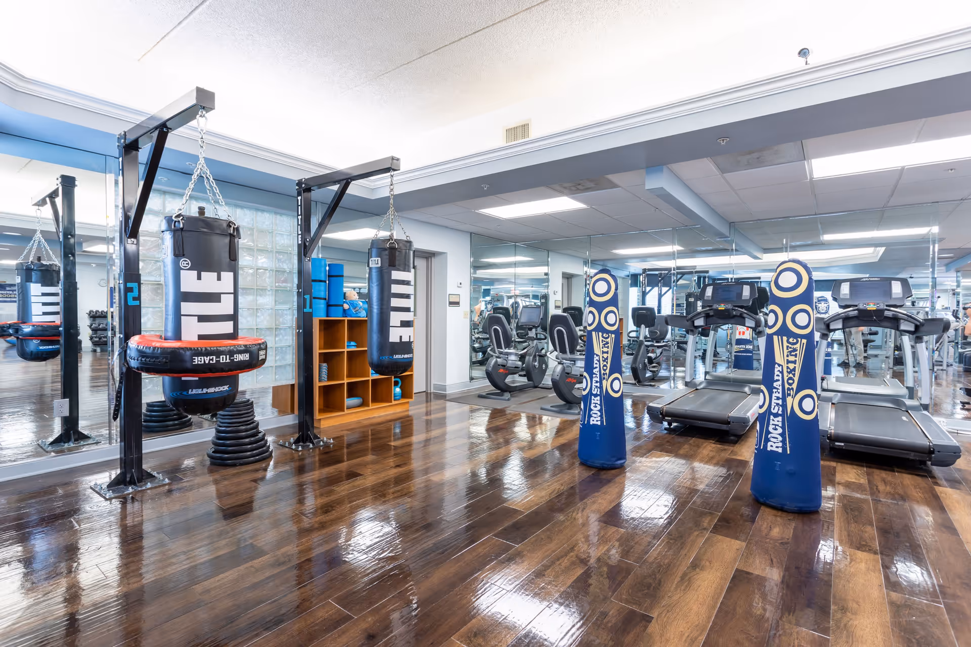 A fitness room with wooden floors featuring two hanging punching bags, two standing punching bags labeled 'Rock Steady Boxing', two treadmills, and exercise bikes. The walls have large mirrors and there is a wooden cubby shelf with blue exercise mats and a teddy bear.
