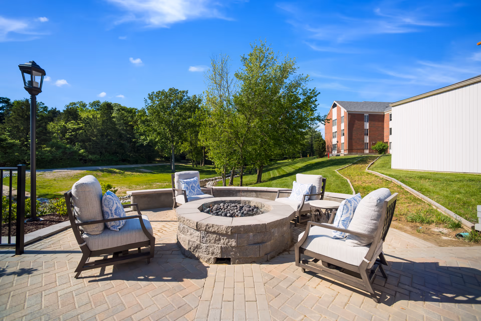 Outdoor patio area with four cushioned chairs arranged around a circular stone fire pit. The patio is paved with light-colored bricks and surrounded by green grass, trees, and a clear blue sky. In the background, there is a multi-story brick building and a white building.
