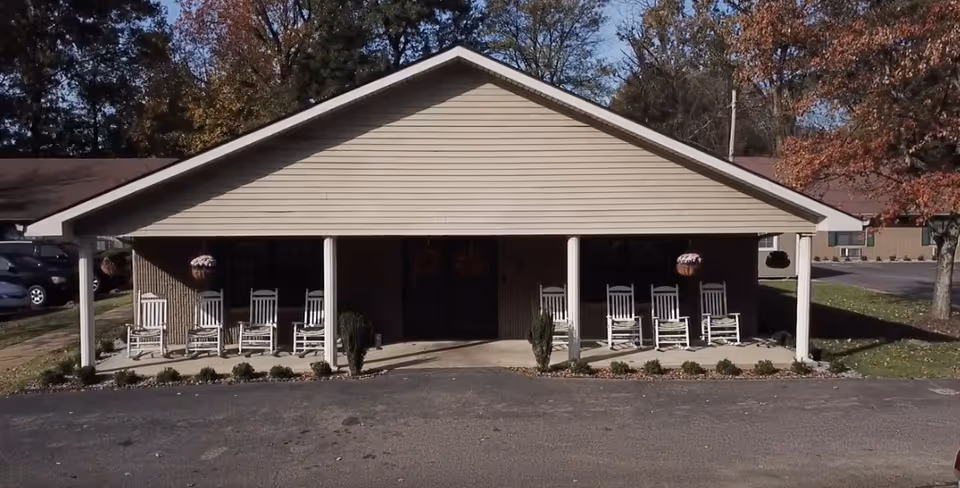 Front entrance of a one-story building with a covered porch lined with white rocking chairs and trees in the background.