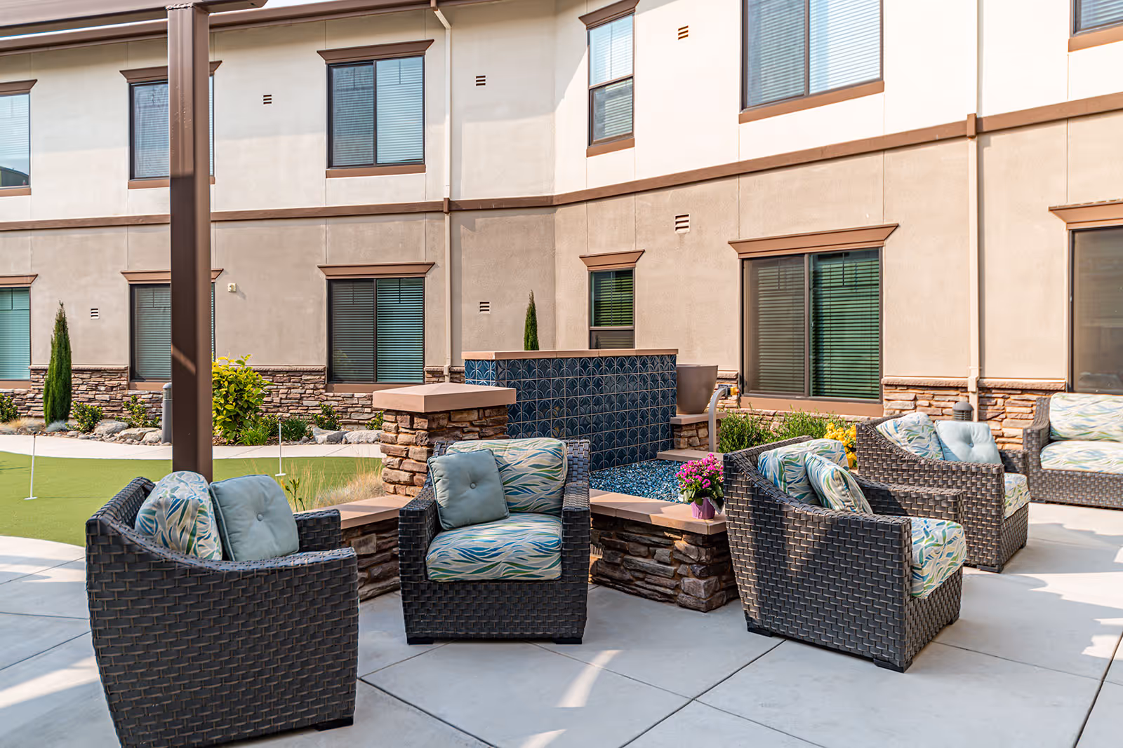 Outdoor patio area at Loma Clara Senior Living featuring several wicker armchairs with patterned cushions arranged around a stone and tile water feature, with a putting green and the building's exterior windows in the background.