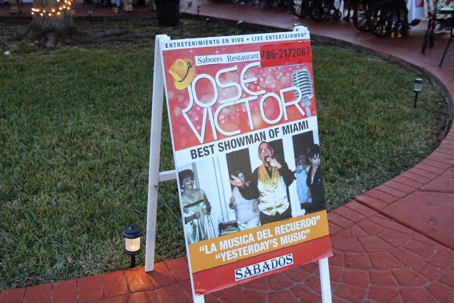 A sidewalk sign on a grassy area advertising live entertainment by Jose Victor, labeled as the best showman of Miami. The sign includes a photo of Jose Victor singing and people enjoying the performance. Text on the sign is in both Spanish and English, promoting 'Yesterday's Music' on Saturdays.