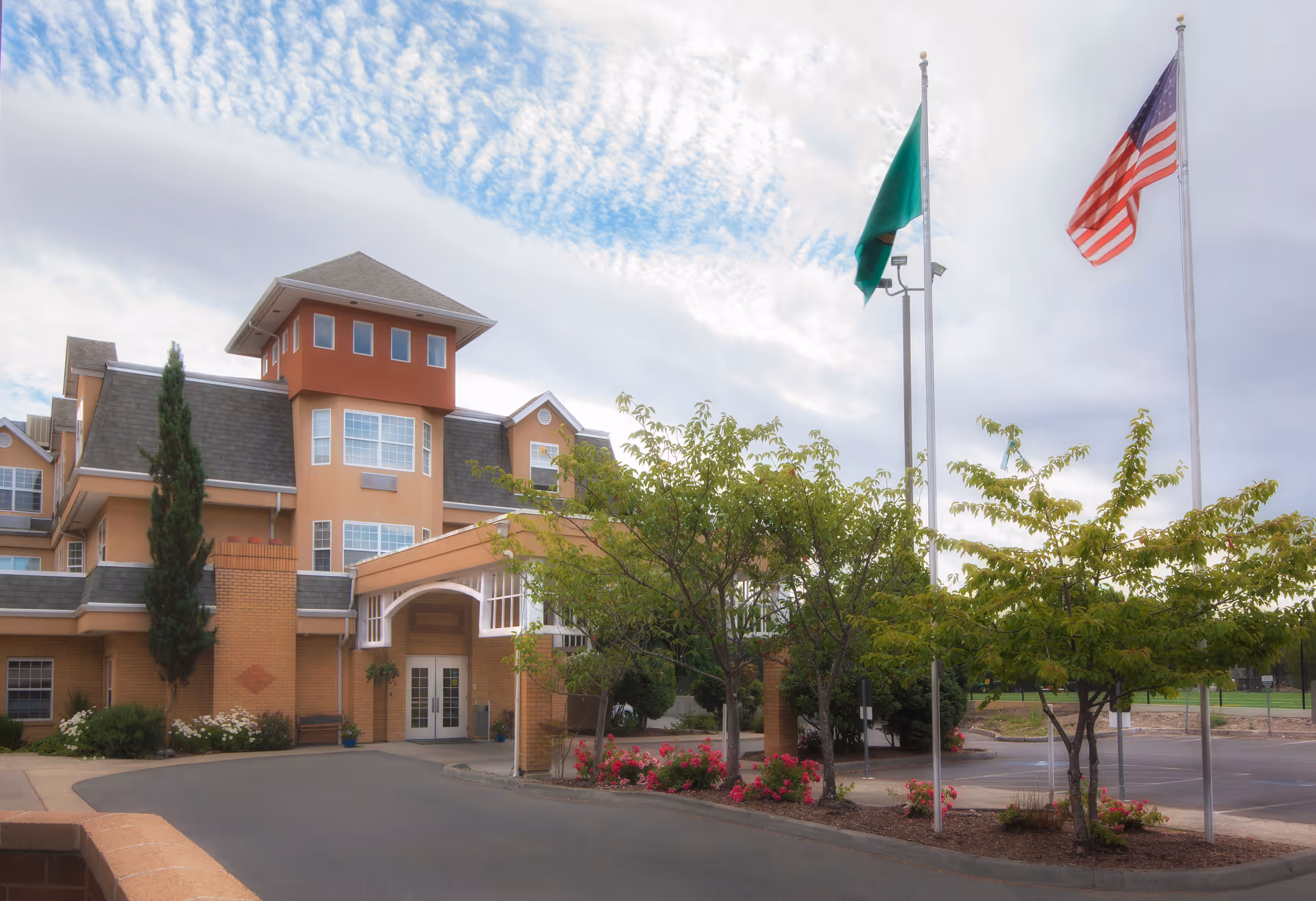 Front entrance of a multi-story senior living building with flags, trees, and landscaped flower beds.