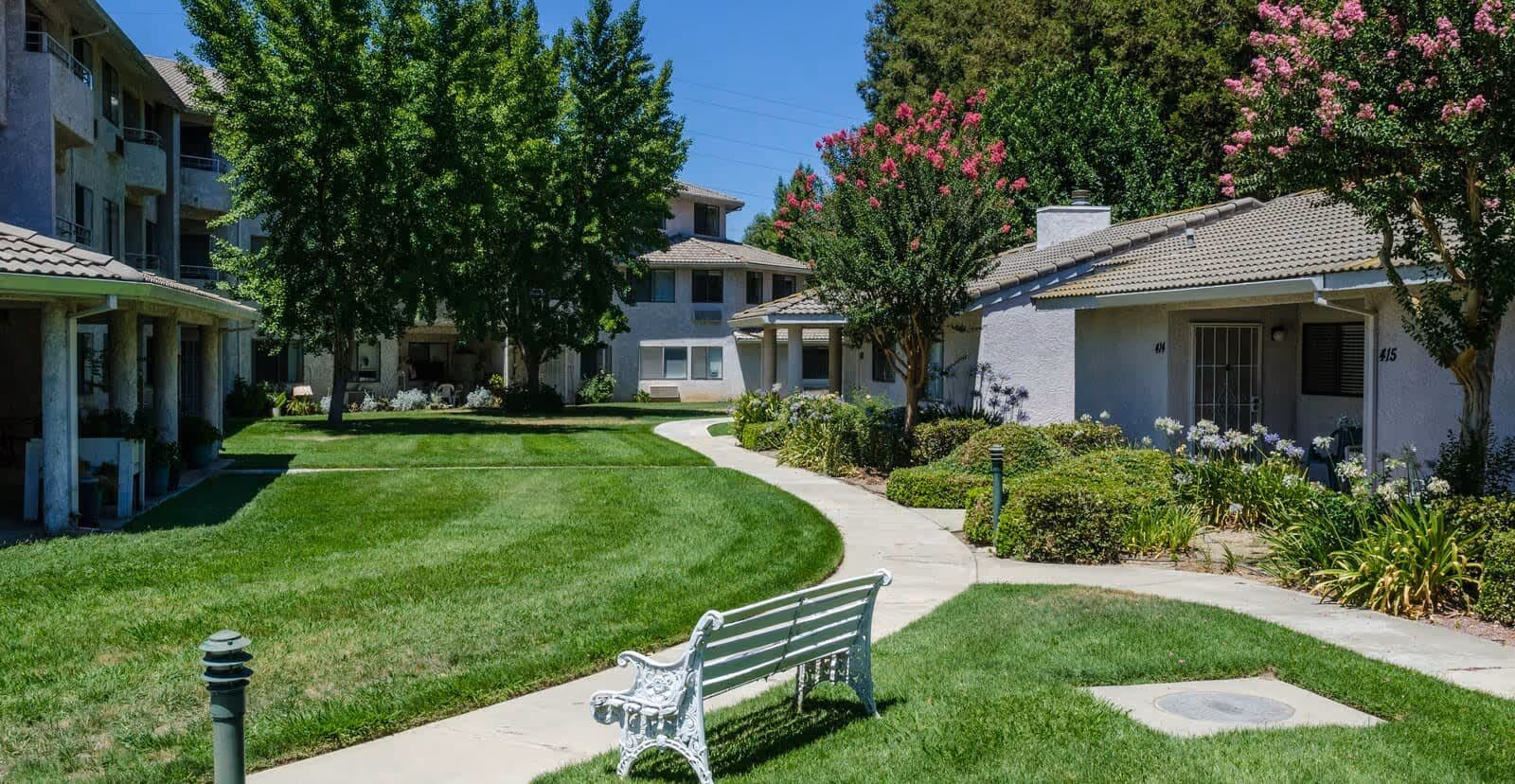 A well-maintained outdoor courtyard area at a senior living facility with green grass, a curved concrete walkway, a white bench, and various trees and shrubs surrounding single-story and multi-story buildings under a clear blue sky.