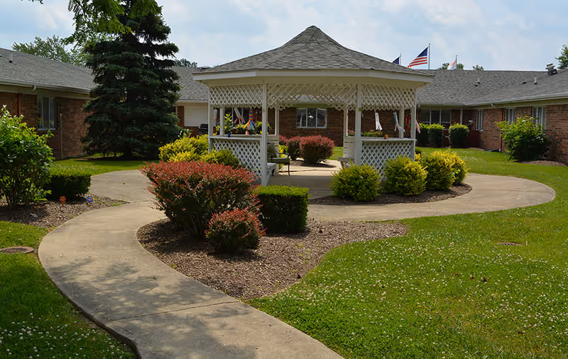 A landscaped courtyard featuring a white gazebo, winding concrete paths, shrubs, and surrounding single-story brick buildings.