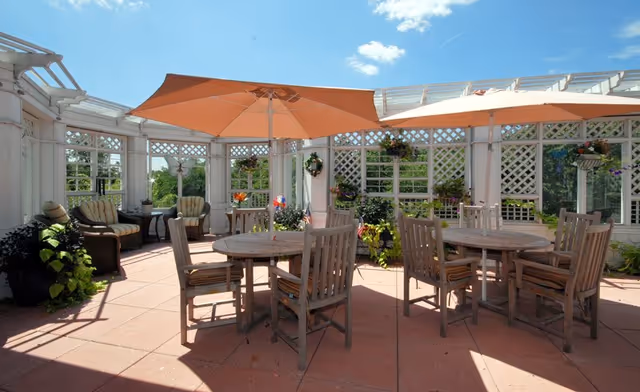 Outdoor patio area with wooden tables and chairs, some tables shaded by large orange umbrellas. The patio is surrounded by white lattice fencing with hanging plants and greenery. There are cushioned armchairs in the corner and a clear blue sky overhead.