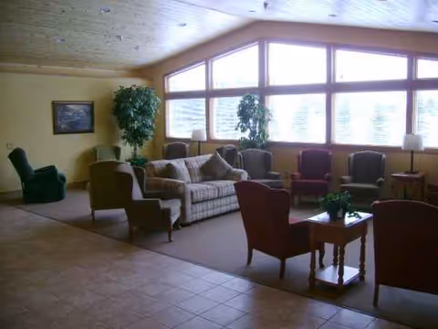 Sunlit senior living lounge with sofas, armchairs, plants, and large front windows.