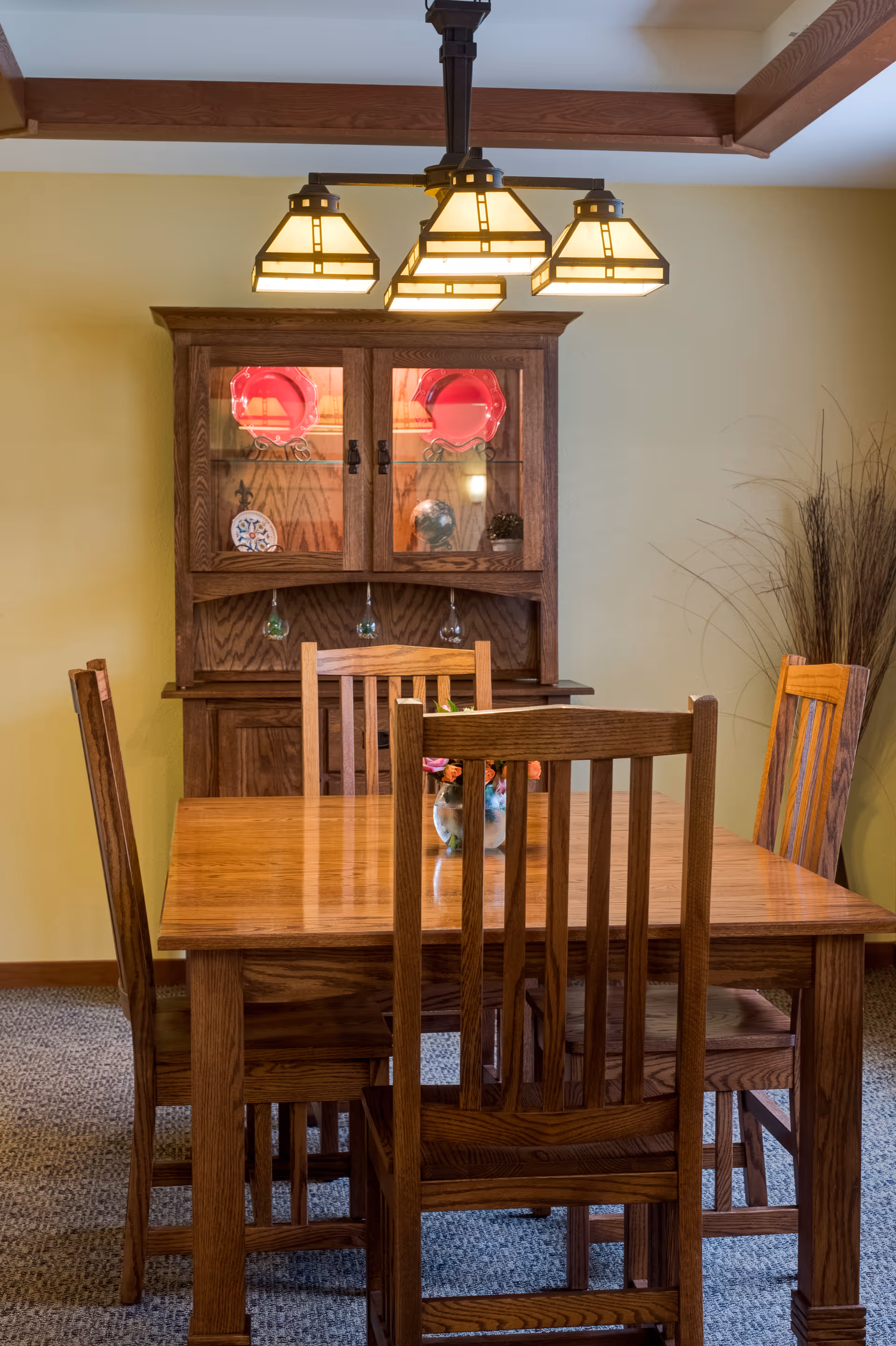 A wooden dining table with four matching wooden chairs in a room with beige walls and carpeted floor. Behind the table is a wooden cabinet with glass doors displaying decorative plates and glassware. Above the table hangs a light fixture with four square lampshades. A tall vase with dried branches is placed in the corner of the room.