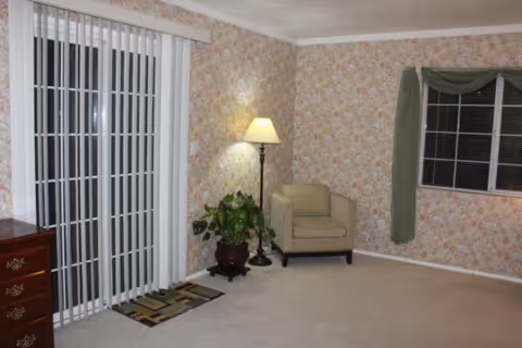 Carpeted living room with floral wallpaper, a single armchair by a floor lamp and potted plant, sliding glass doors with vertical blinds, and a window with a green valance.