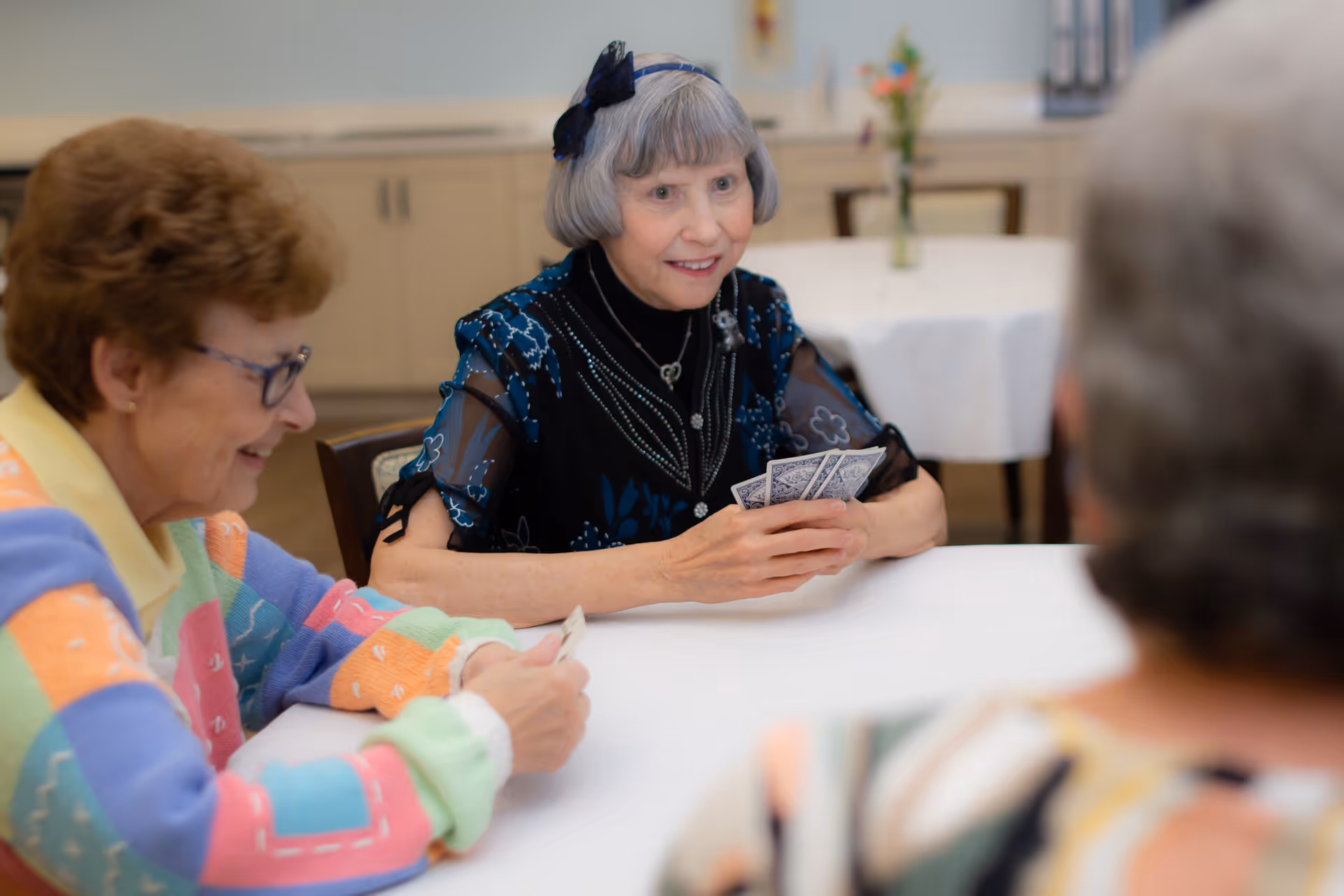 Three elderly women sitting around a table playing cards in a well-lit room with cabinets and a table with a vase of flowers in the background.