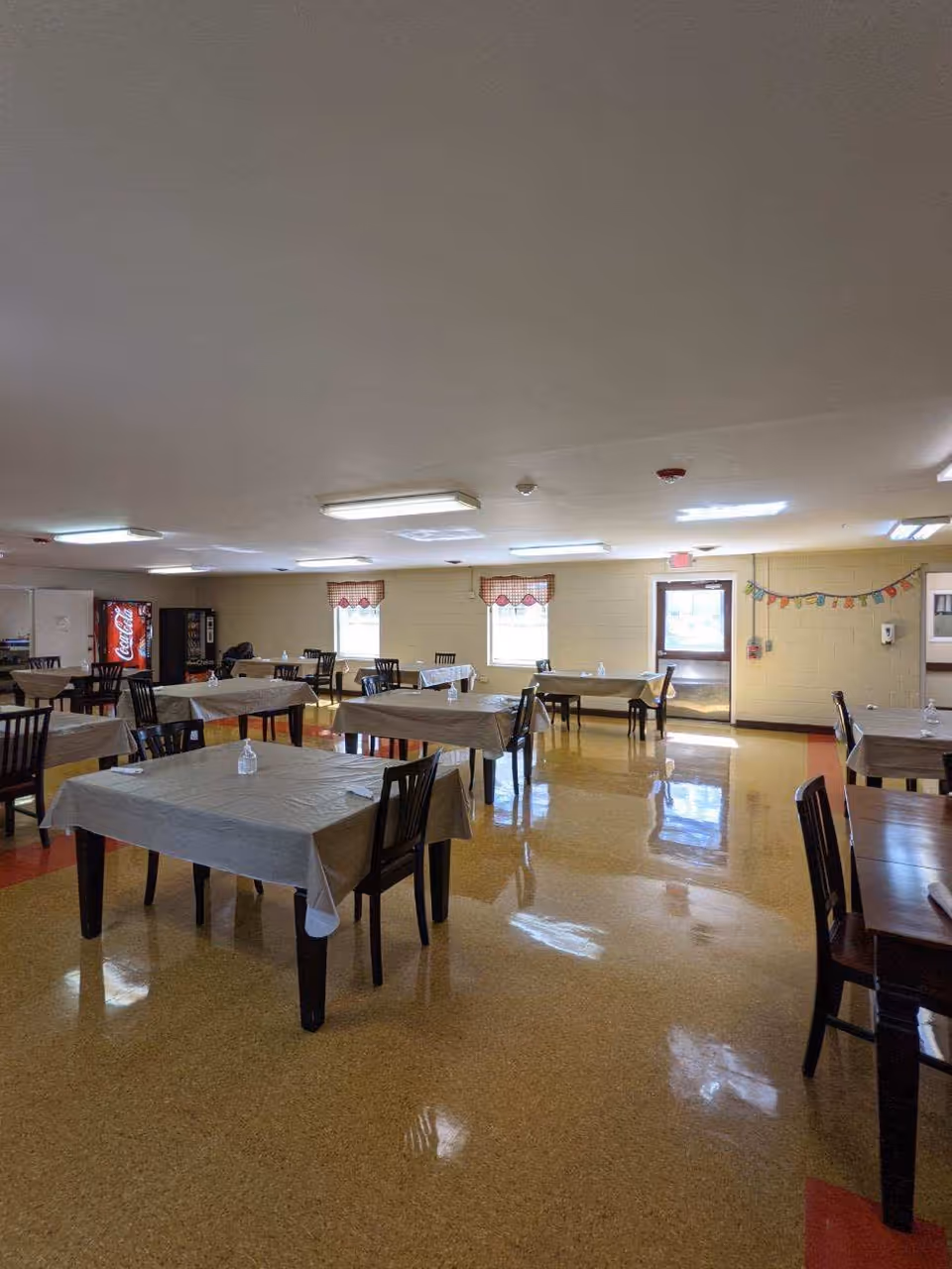 A dining room with multiple tables covered with beige tablecloths and surrounded by dark wooden chairs. The room has a shiny yellow and red floor, beige walls, and several fluorescent ceiling lights. There are two windows with red and white checkered valances, a door with a window, and a Coca-Cola vending machine in the corner.