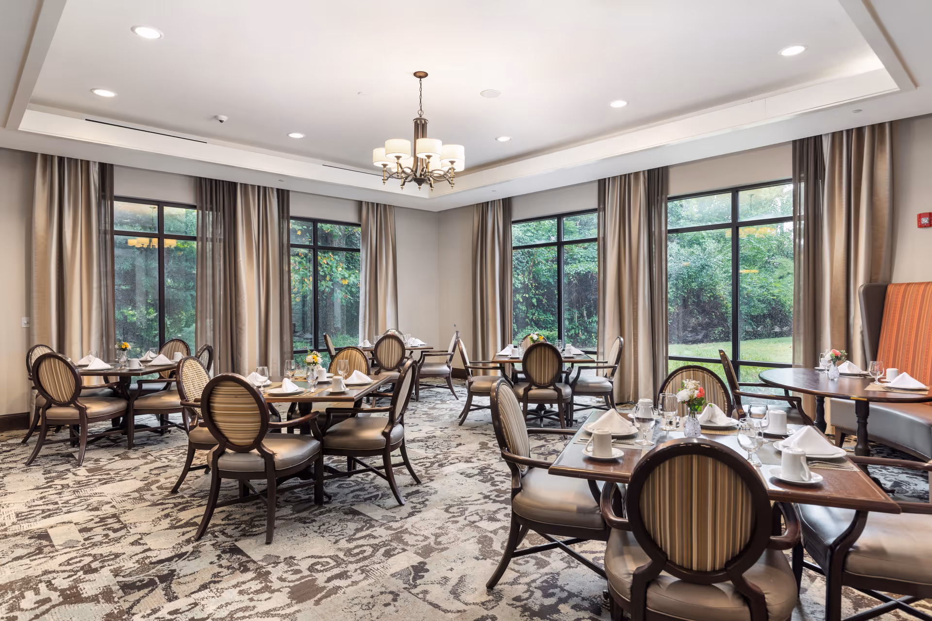 A bright and elegant dining room with several round wooden tables set with white napkins, cups, glasses, and small flower arrangements. The room has large windows with beige curtains, allowing natural light to illuminate the space. The floor is covered with a patterned carpet, and a chandelier hangs from the ceiling.