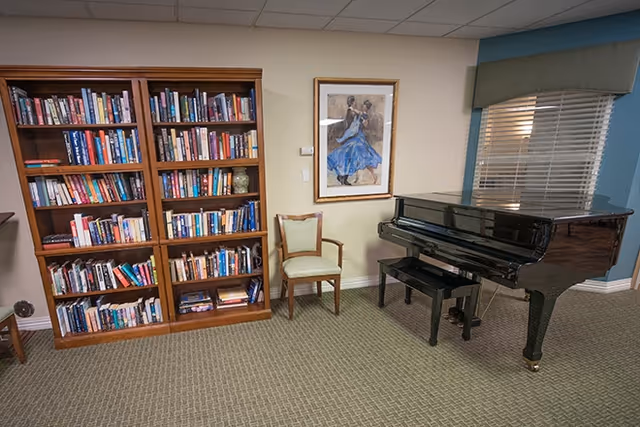 A common room with a large wooden bookshelf, a black grand piano, a chair, framed artwork, and a window with blinds.