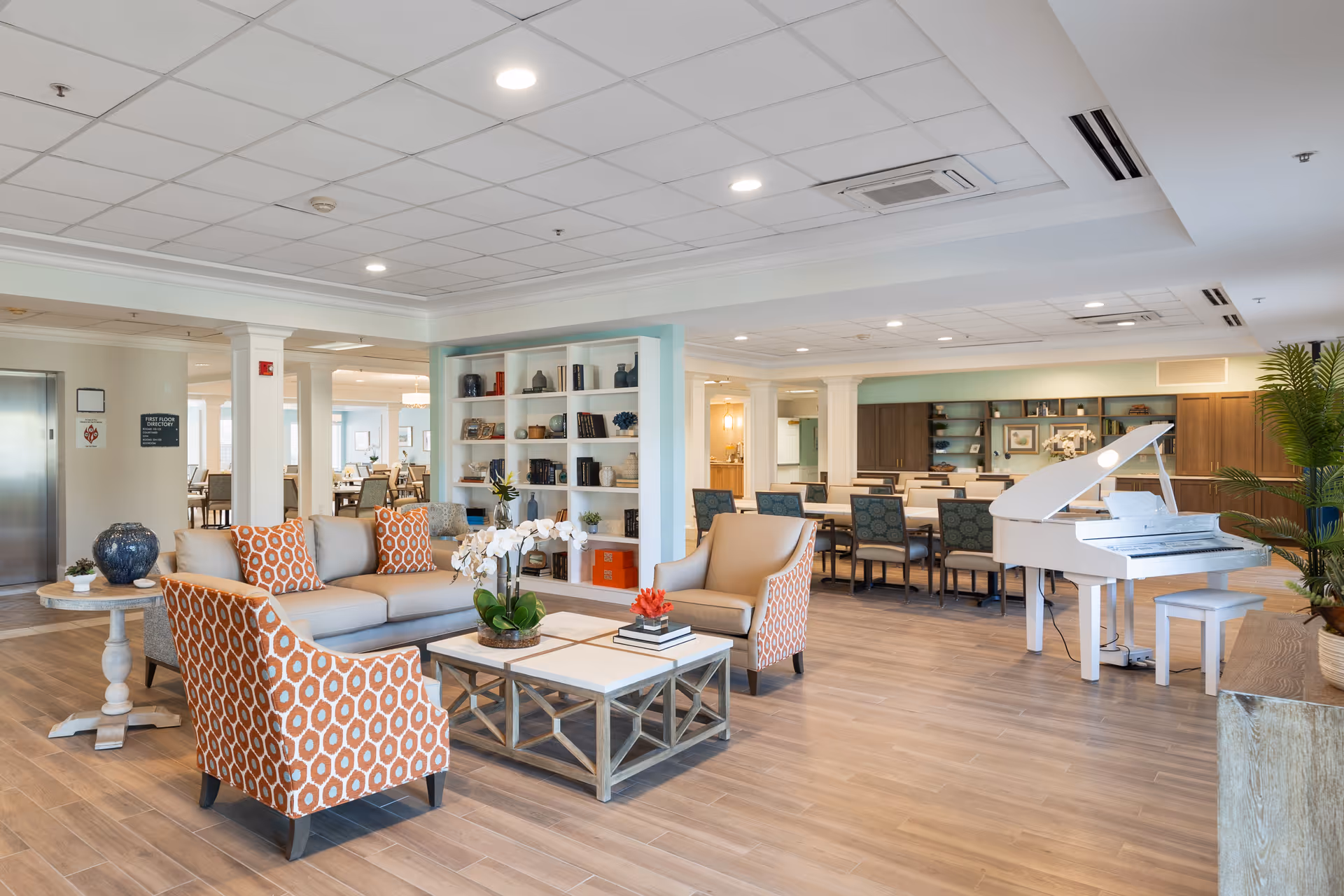 A spacious and well-lit senior living facility common area featuring a seating arrangement with a beige sofa, two orange patterned armchairs, a coffee table with decorative items, a white grand piano, and a dining area with tables and chairs in the background. The room has wooden flooring, built-in shelves with decorative items, and a modern ceiling with recessed lighting.