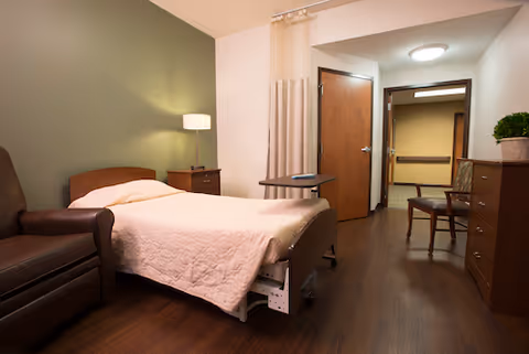 A senior living bedroom with an adjustable bed, armchair, nightstand with lamp, dresser, and a doorway leading to a hallway.