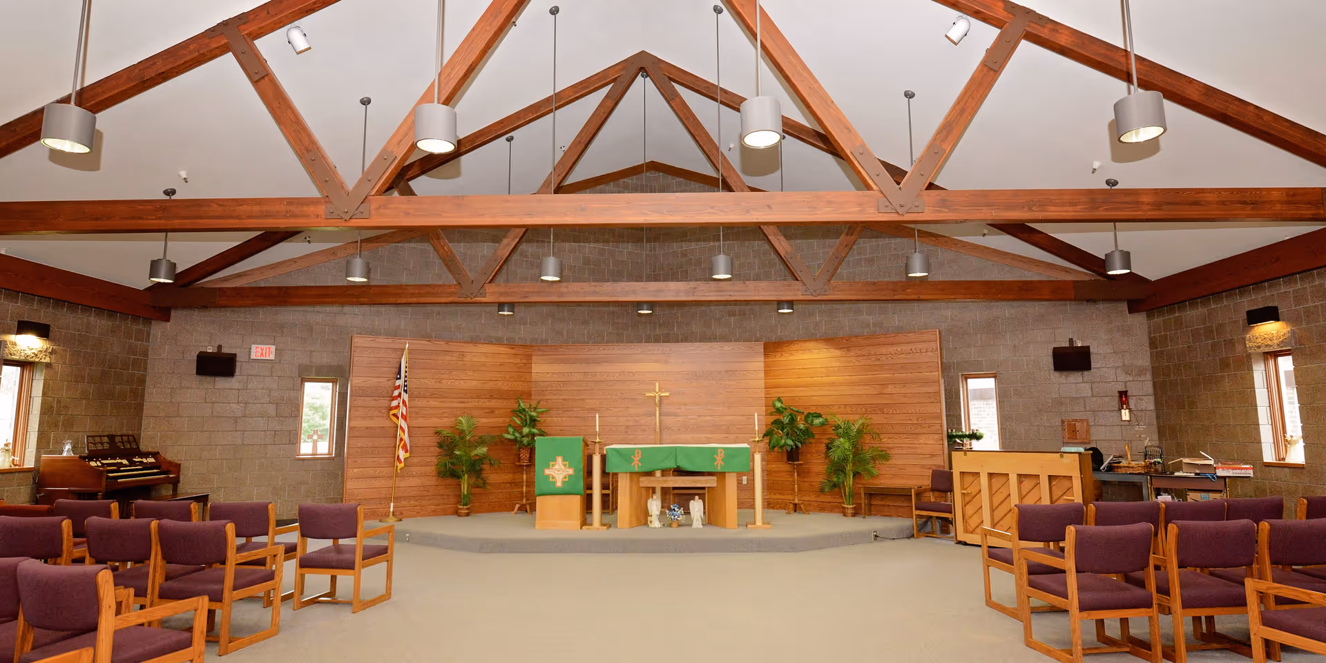 Interior chapel with exposed wooden ceiling trusses, rows of purple chairs facing an altar with a cross, lectern, piano and an American flag.