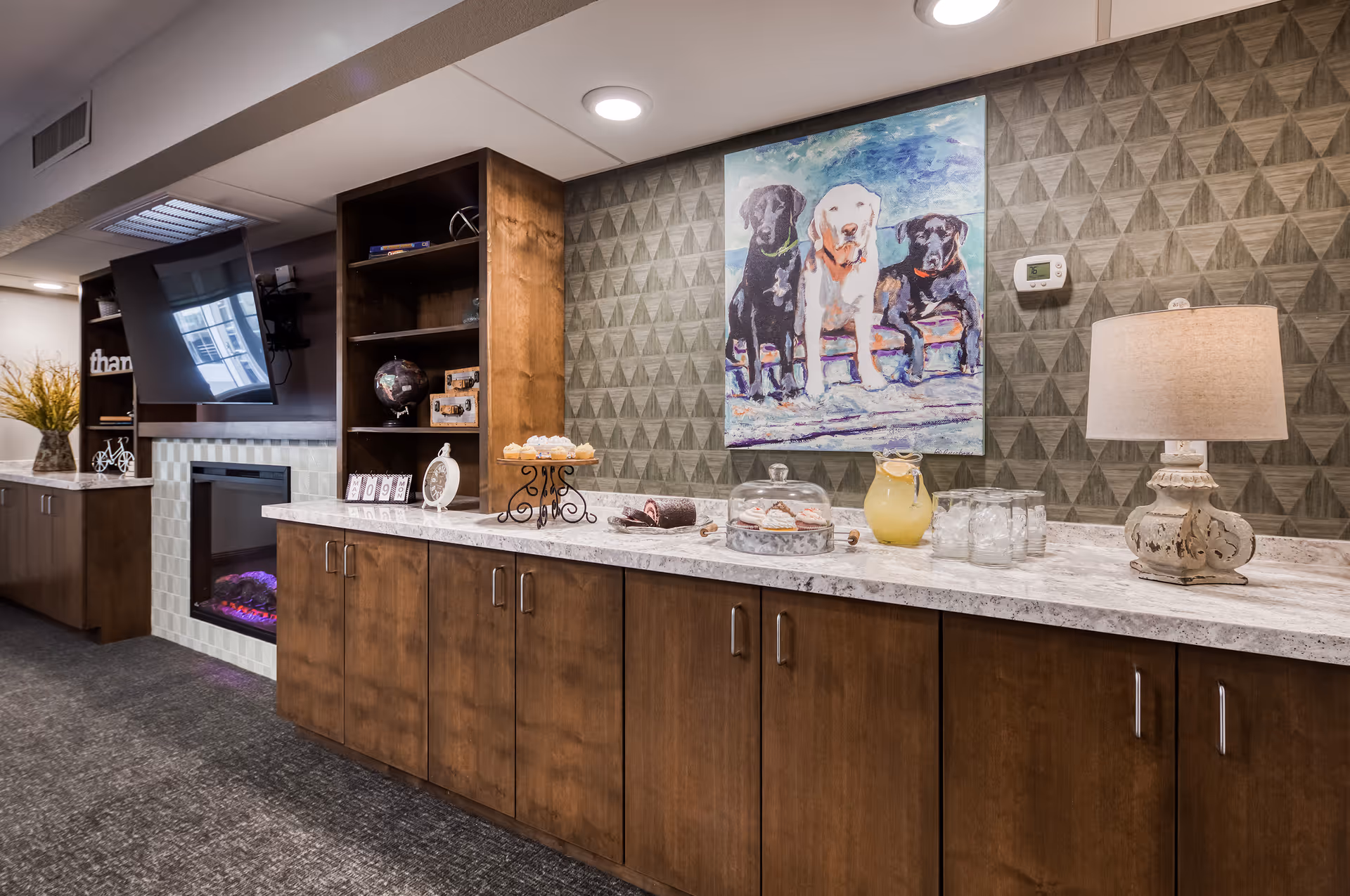 Interior view of a common area with a long wooden cabinet topped with a marble countertop. On the countertop are cupcakes, a cake roll, a glass dome with pastries, a pitcher of lemonade with glasses, and a decorative lamp. Above the cabinet is a painting of three dogs on a textured wall. To the left, there is a mounted flat-screen TV above an electric fireplace and built-in shelves with decorative items.