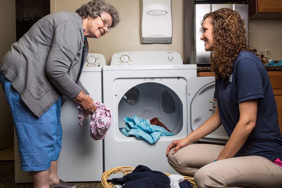 An elderly woman and a younger woman are doing laundry together. The elderly woman is standing and holding clothes near a washing machine, while the younger woman is kneeling beside an open dryer with clothes inside. They are in a laundry room with cabinets and a paper towel dispenser on the wall.