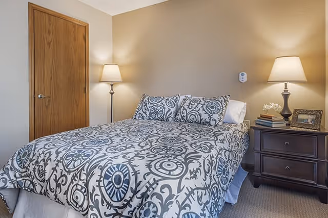 A bedroom featuring a bed with a patterned gray and white bedspread and matching pillows. There is a wooden door on the left side, a nightstand on the right with a lamp, books, a small flower arrangement, and a framed photo. The walls are beige and the floor is carpeted.