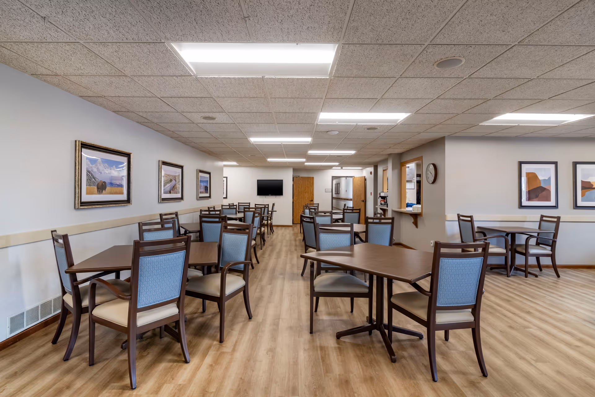A spacious dining room in a senior living facility with multiple wooden tables and chairs arranged neatly. The room has light-colored walls adorned with framed landscape artwork, a wood floor, and a drop ceiling with fluorescent lighting. A wall-mounted TV is visible at the far end, and there is a service window on the right side with a clock above it.