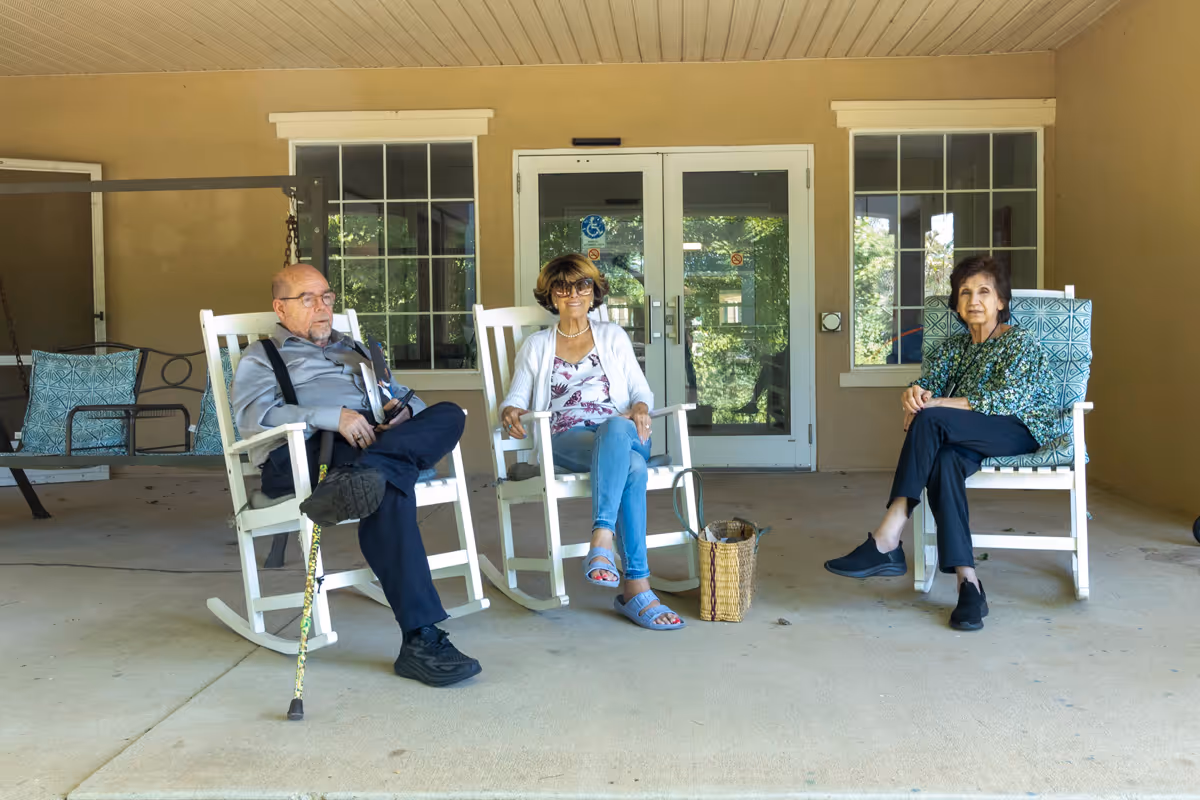 Three elderly individuals sitting on white rocking chairs on a covered porch area outside a building. The man on the left has a cane resting against his chair, the woman in the middle is wearing sunglasses and casual clothes, and the woman on the right is seated comfortably with her legs crossed. Behind them are two windows and a glass door with reflections of greenery outside.
