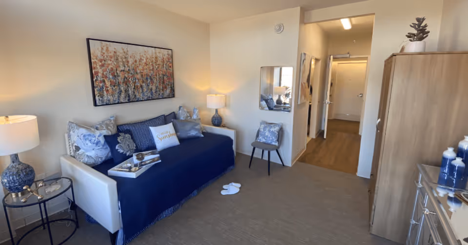 A cozy living room area in a senior living facility with a white sofa covered by a dark blue throw and multiple decorative pillows, including one that says 'Hello Sunshine.' There is a side table with a blue lamp and decorative items, a chair with a floral cushion, a wooden cabinet, and a hallway leading to another room. The walls are light-colored, and a large abstract painting hangs above the sofa.