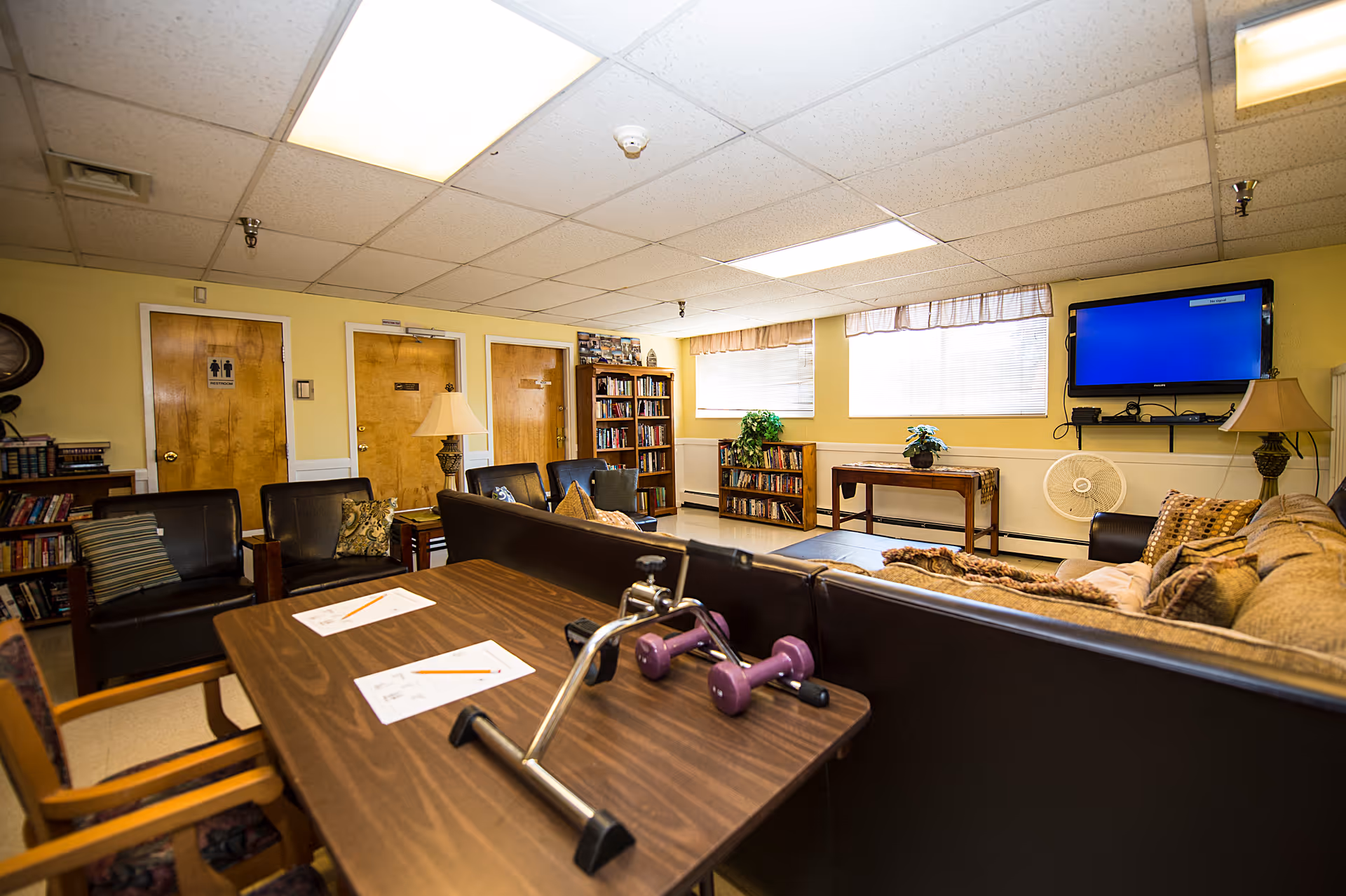 A cozy common area in a senior living facility with comfortable seating including a sofa and armchairs, a wooden table with exercise equipment and papers on it, bookshelves filled with books, a wall-mounted TV, and windows letting in natural light. The walls are painted yellow and there are wooden doors in the background.