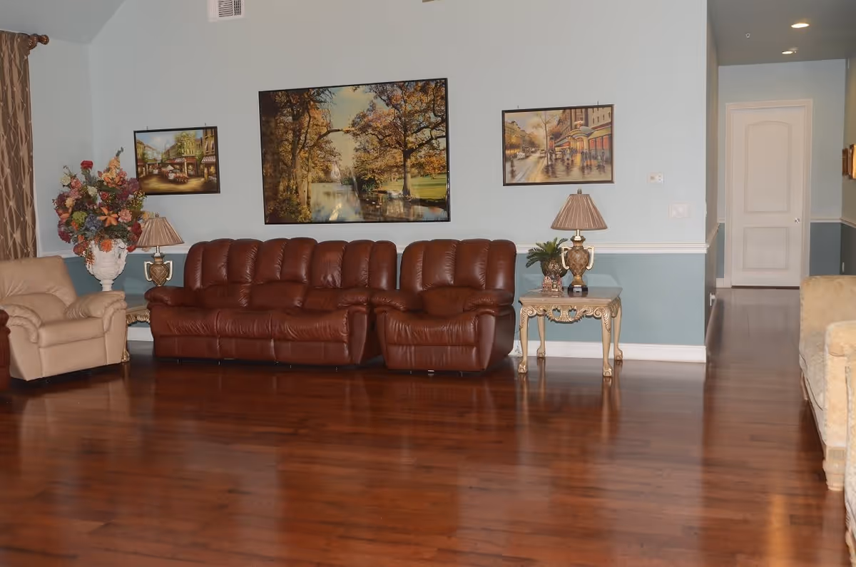 A living room with a brown leather sofa and a beige armchair on a polished wooden floor. There are three framed paintings on the light blue wall above the sofa. Two ornate side tables with decorative lamps and a large floral arrangement are placed on either side of the sofa. A hallway with a white door is visible in the background.