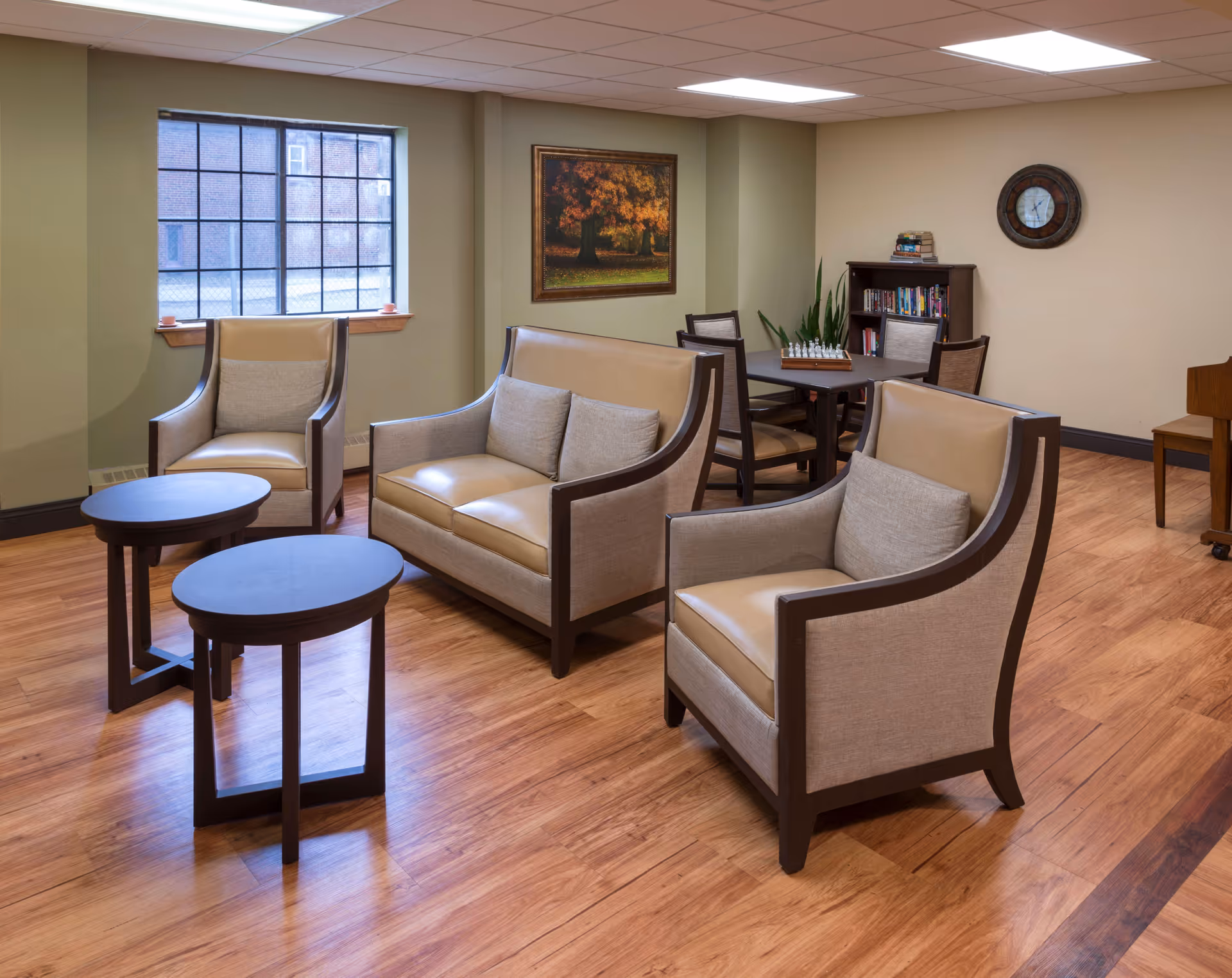 A cozy sitting area in a senior living facility with two beige armchairs and a matching loveseat arranged around two small round wooden tables. In the background, there is a table with four chairs, a bookshelf with books and a chess set, a wall clock, and a framed painting of a tree with autumn leaves. The room has wooden flooring and a window letting in natural light.