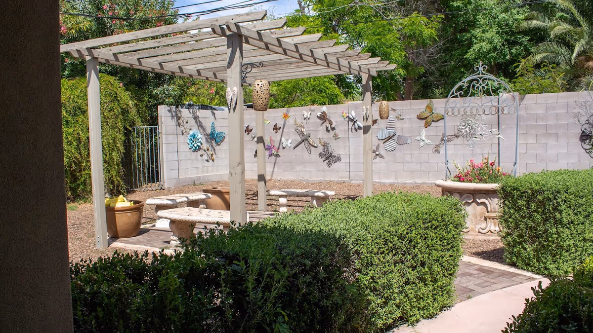 Outdoor garden area with a wooden pergola, stone benches and tables, decorative metal butterflies and dragonflies on a concrete block wall, potted plants, and trimmed green bushes.