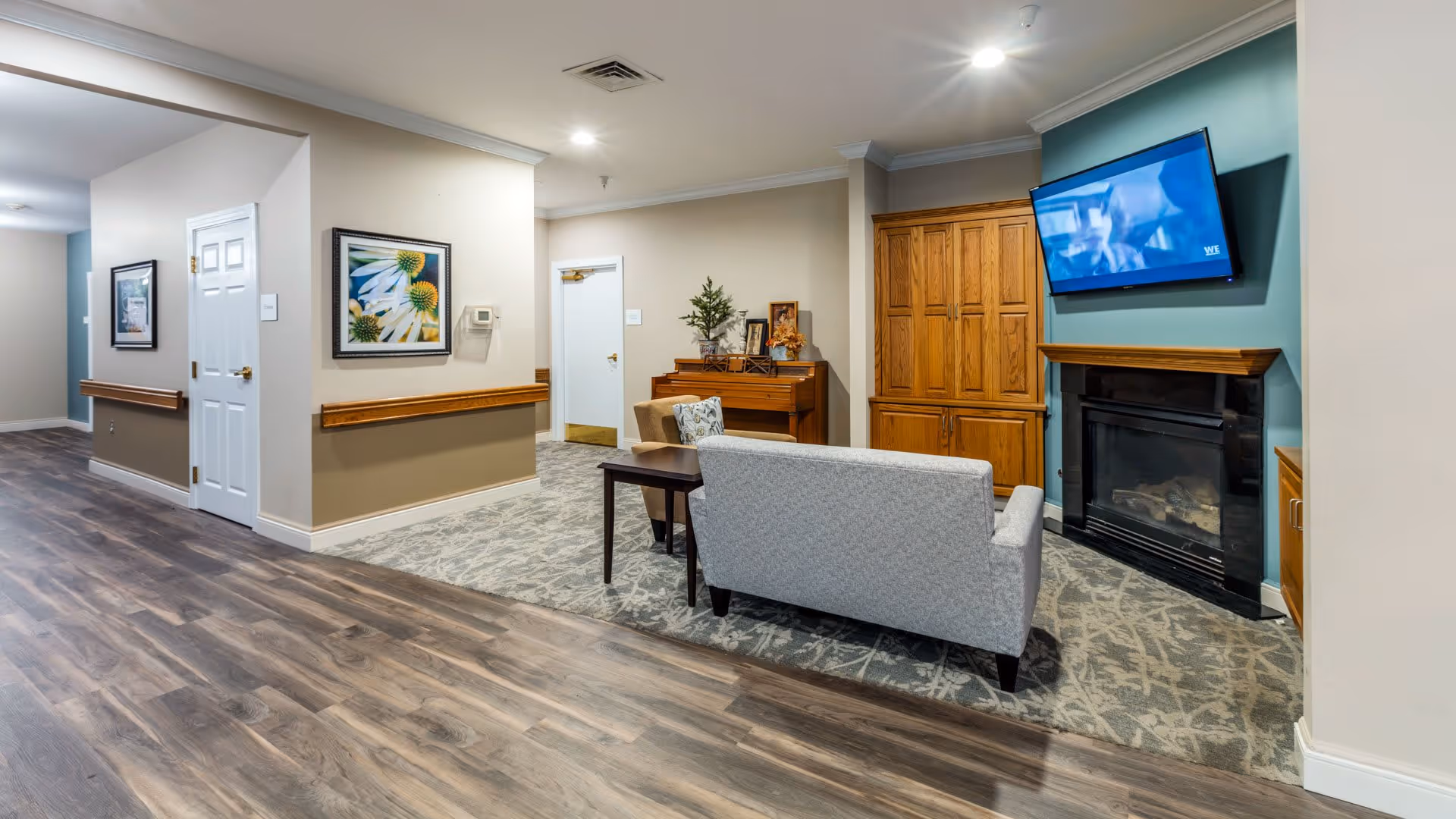 A cozy common area in a senior living facility featuring a light gray couch facing a wall-mounted flat screen TV above a fireplace. There is a wooden cabinet and a piano with decorative items on top. The floor has a mix of wood and carpet, and the walls are painted in neutral tones with framed artwork.