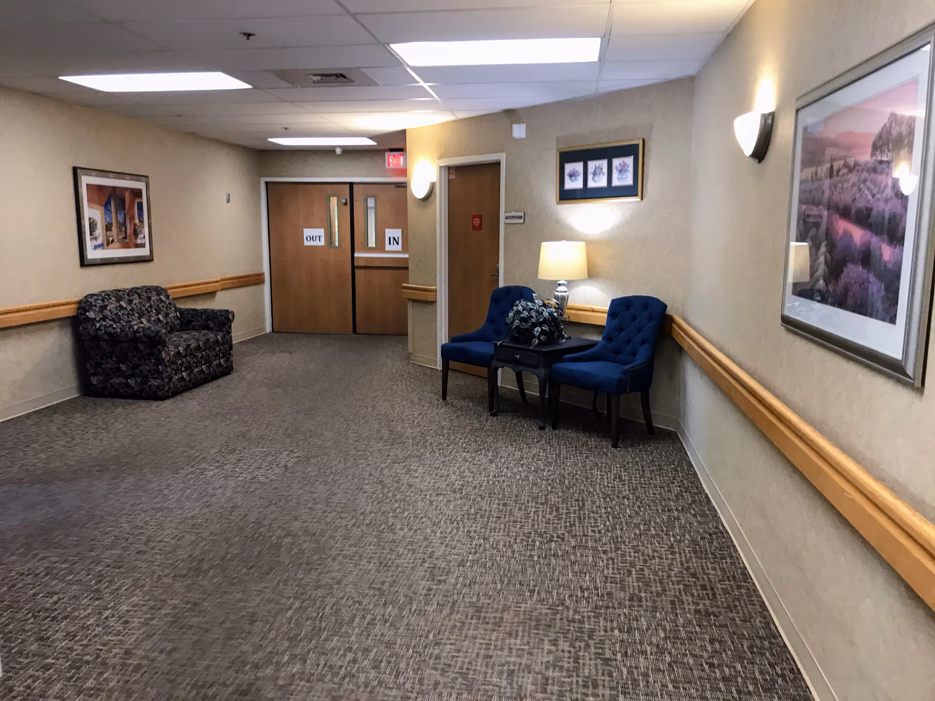 A hallway in a senior living facility with beige walls and carpeted floor. There are two blue upholstered chairs with a small table and lamp between them on the right side, and a floral-patterned loveseat on the left side. Framed artwork hangs on the walls, and double wooden doors labeled 'IN' and 'OUT' are visible at the end of the hallway.