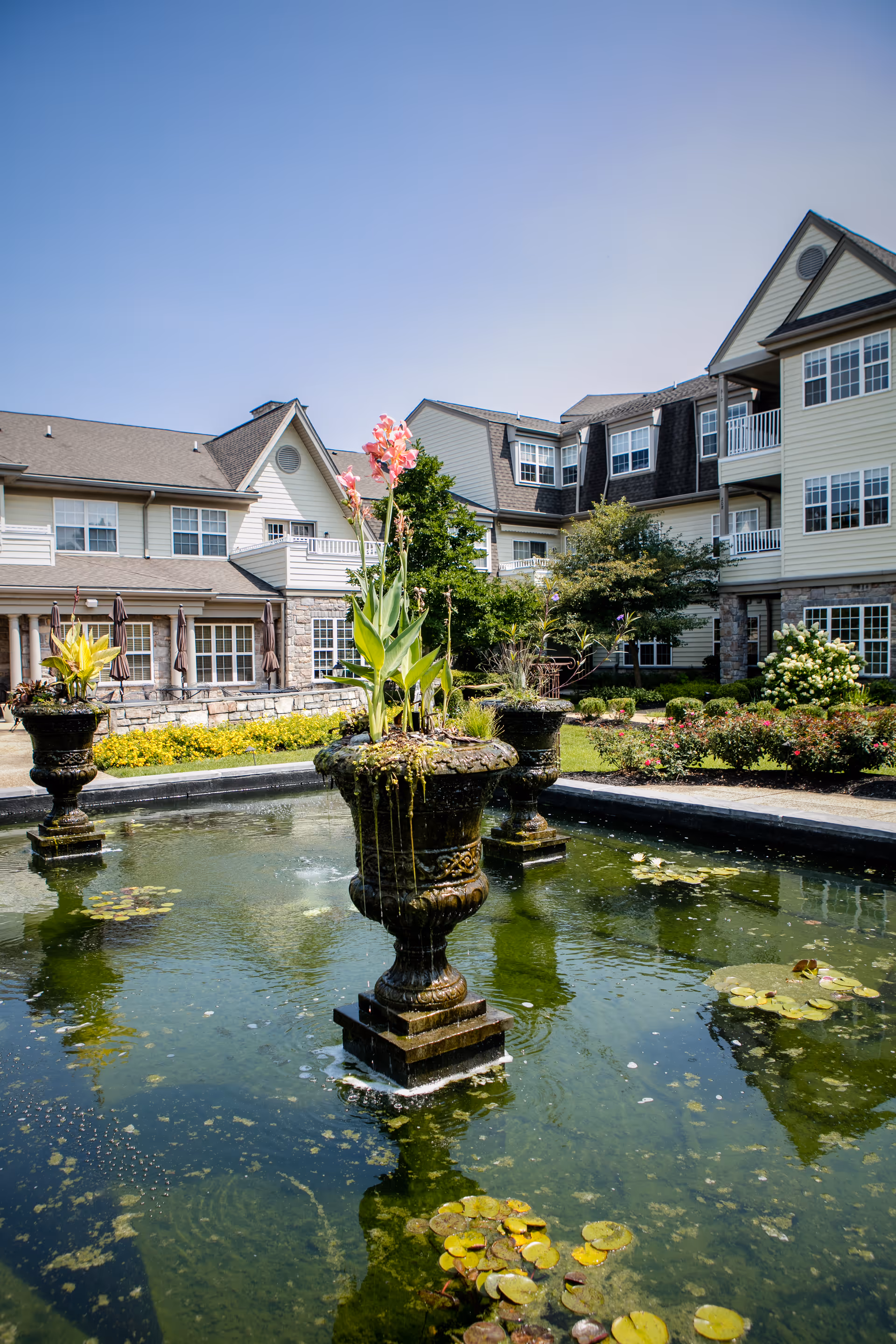 Outdoor courtyard area of a senior living facility featuring a decorative water fountain with plants in the center of a pond surrounded by lily pads. The background shows a multi-story building with balconies, windows, and well-maintained landscaping under a clear blue sky.