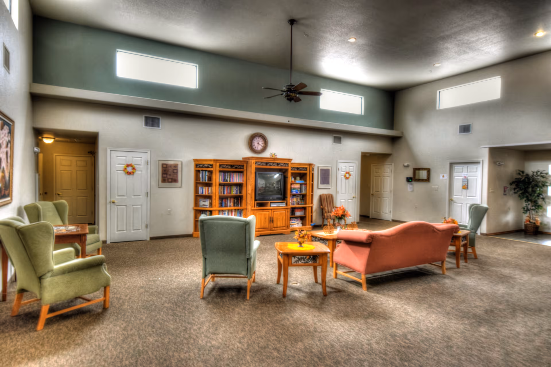 A spacious senior living facility common area with multiple armchairs and a sofa arranged around wooden coffee tables. A wooden entertainment center with a TV and bookshelves is against the far wall, which also has a clock mounted above it. The room has high ceilings with three rectangular windows near the ceiling and a ceiling fan in the center. Several doors and a potted plant are visible along the walls.