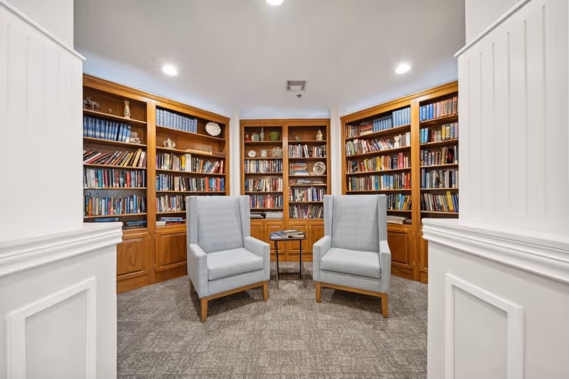 A cozy library room with wooden bookshelves filled with books and decorative items. Two light gray upholstered armchairs face each other with a small round table between them. The room has white paneled walls and a carpeted floor.