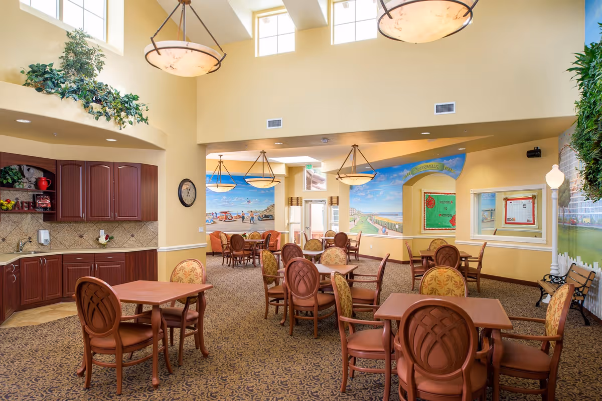 A bright and spacious dining room with multiple wooden tables and chairs arranged neatly. The room features high ceilings with large hanging light fixtures and several windows letting in natural light. There is a kitchenette area with cabinets and a sink on the left side. The walls are decorated with murals depicting outdoor scenes and a bulletin board. The carpet has a patterned design, and there are some plants adding greenery to the space.