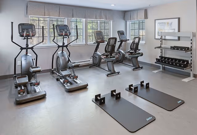 A fitness room with exercise equipment including two elliptical machines, two recumbent stationary bikes, two exercise mats with small dumbbells on them, and a rack of various dumbbells. The room has large windows with gray valances and light gray walls.