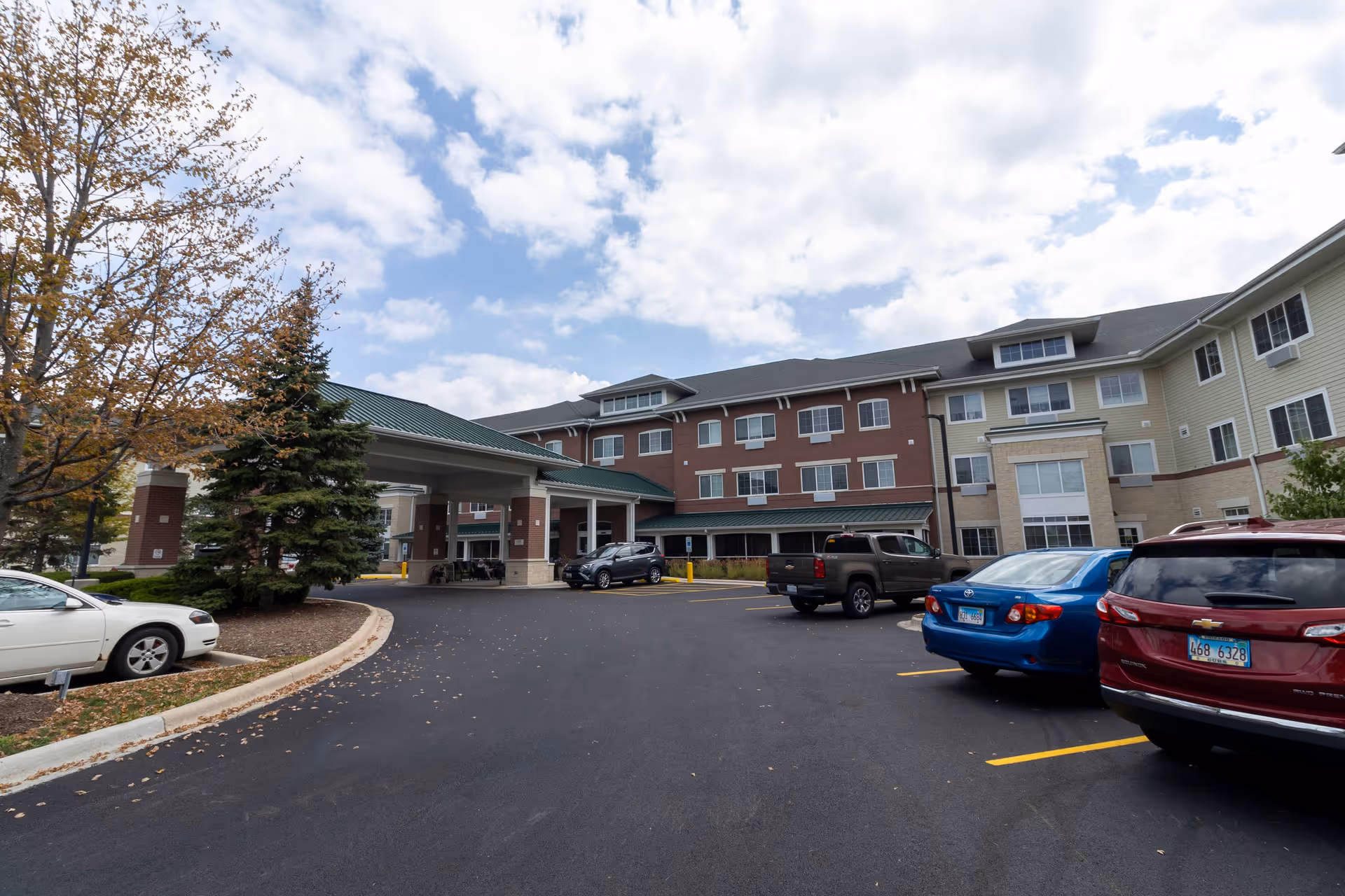 Exterior view of a multi-story senior living facility with a covered entrance and several parked cars in the parking lot under a partly cloudy sky.