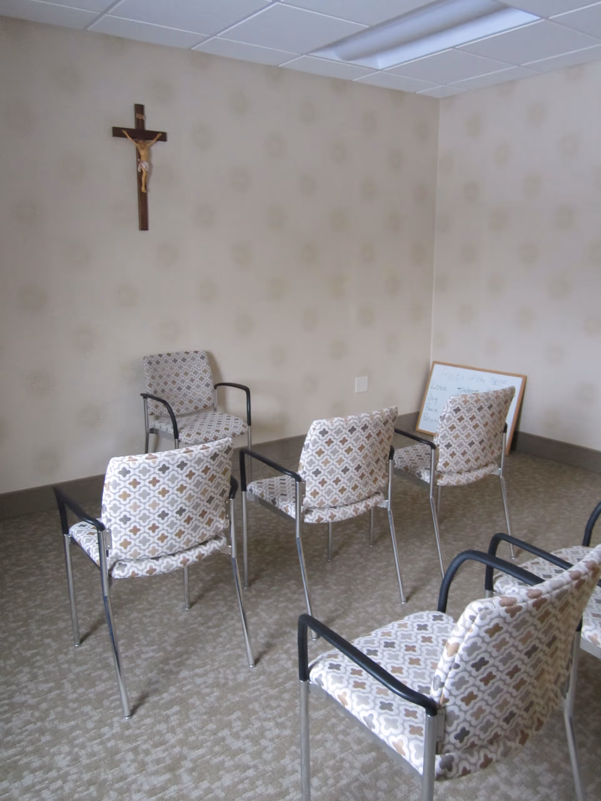 Small interior meeting/chapel room with patterned chairs arranged in rows, a crucifix on the wall and a framed sign leaning in the corner.