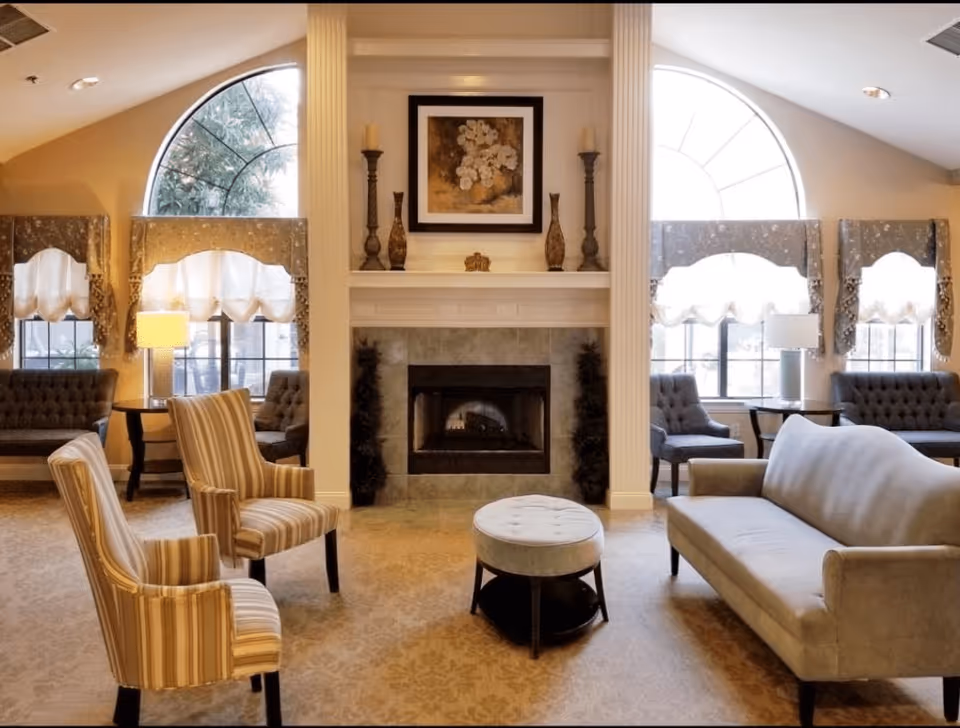 A cozy living room area with a central fireplace flanked by two large arched windows with decorative curtains. The room features a beige sofa, two striped armchairs, a round cushioned ottoman, and additional seating along the walls. The fireplace mantel is decorated with candlesticks, vases, and a framed floral painting above it. The room is softly lit with table lamps and natural light from the windows.