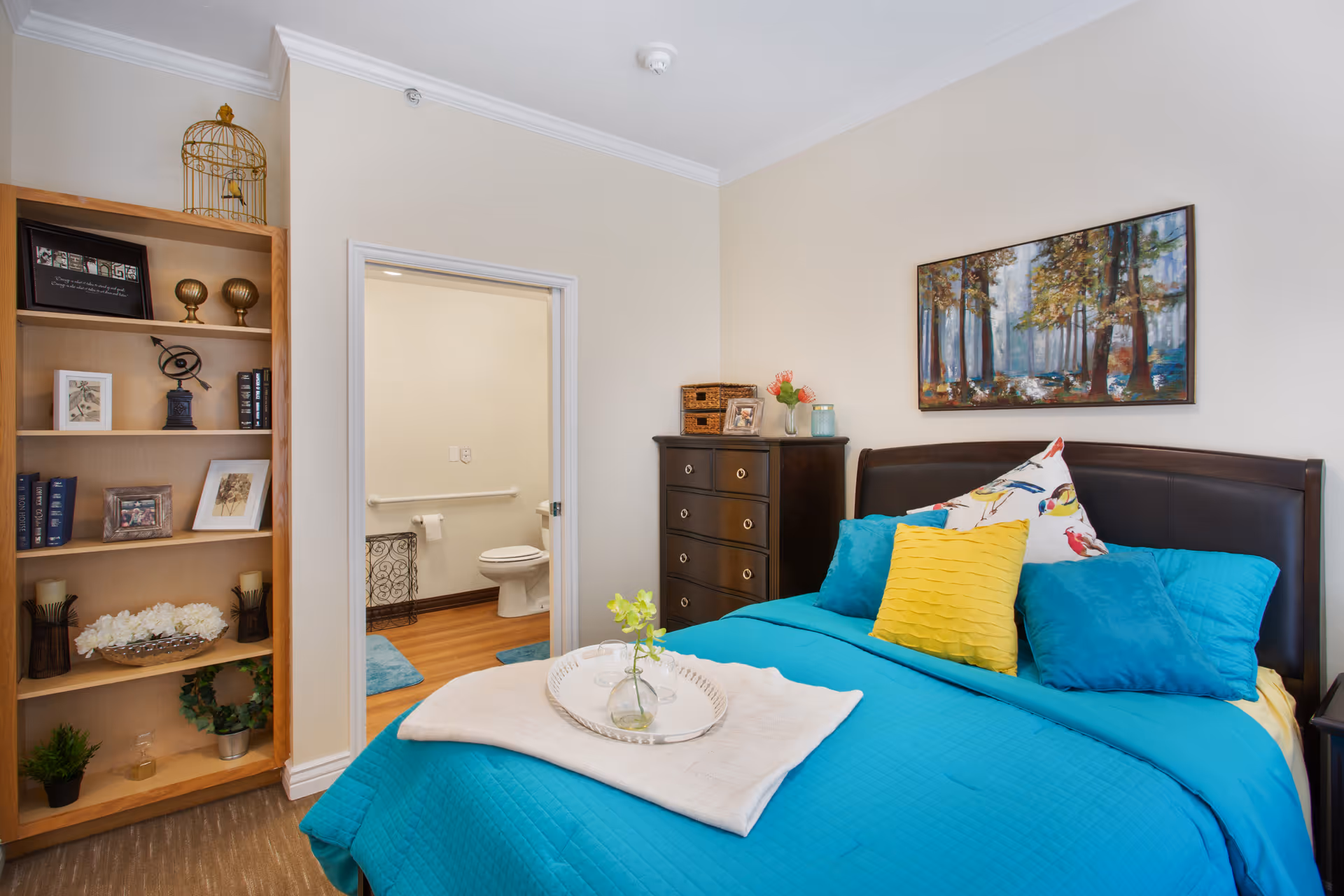 A cozy bedroom with a bed dressed in bright blue bedding and colorful pillows, a dark wooden dresser, and a bookshelf filled with decorative items. A doorway leads to a bathroom with a visible toilet and grab bars. A framed painting of a forest hangs above the bed.